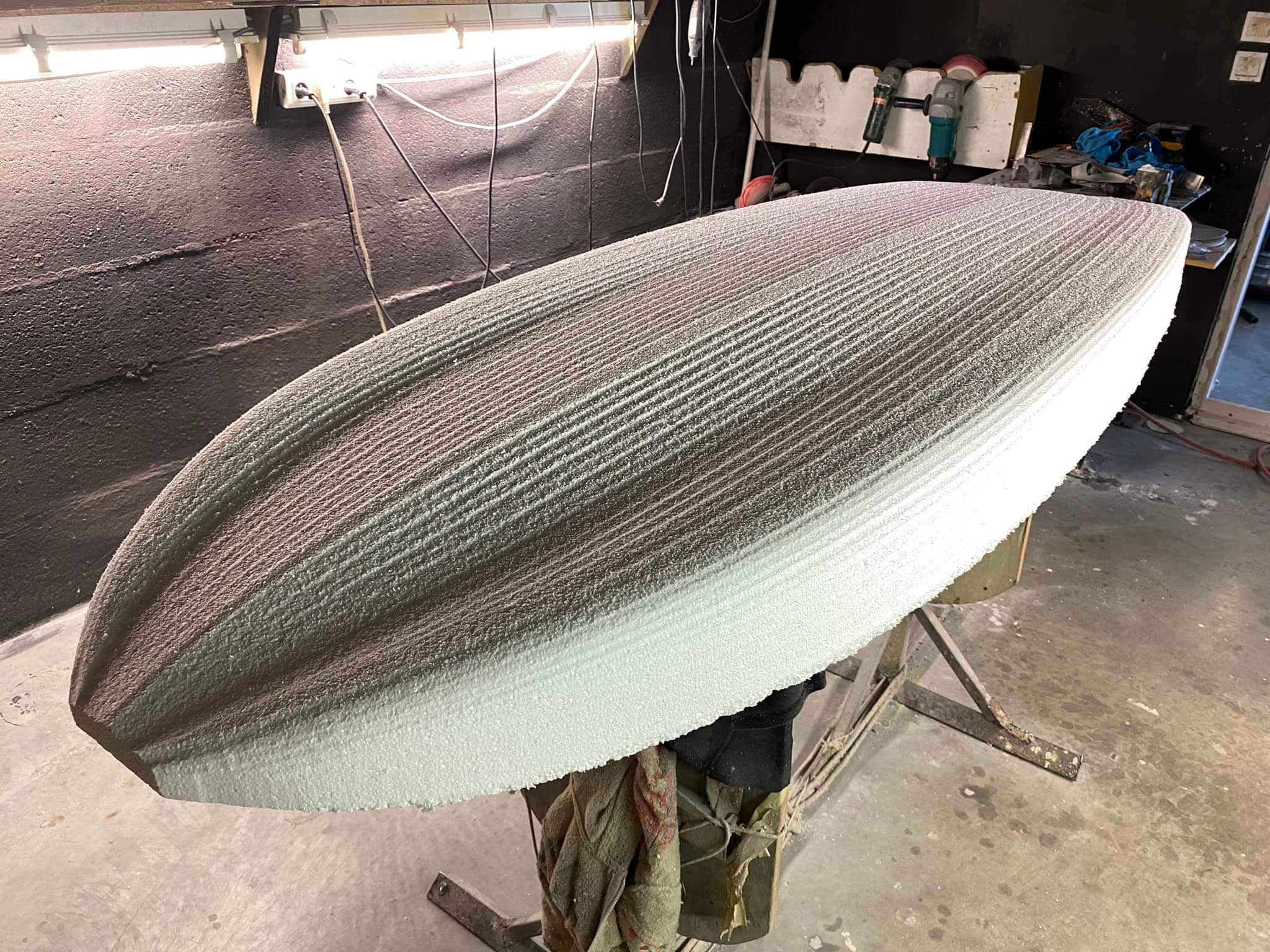 Une grande planche de surf en mousse en cours de construction sur un support dans un atelier ressemble à une planche de SUP. La planche a une surface texturée avec des rainures, et les outils et l'équipement sont dispersés sur le mur sombre à l'arrière-plan.