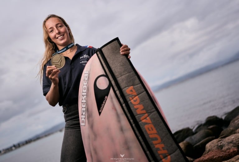 Une femme souriante, tenant une médaille et un bodyboard, se tient debout sur les rochers au bord de la mer à La Rochelle. Elle porte une chemise bleu marine avec des logos, incarnant une silhouette confiante. Le ciel est couvert en arrière-plan, mettant en valeur la scène côtière spectaculaire.