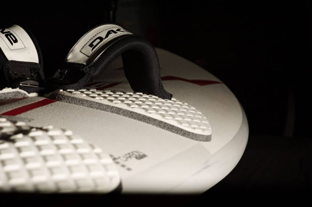 Gros plan d'une planche de surf blanche avec des repose-pieds texturés et des sangles de pied noires étiquetées « DAKINE » sur un fond sombre. La planche présente des accents et un texte rouges subtils.