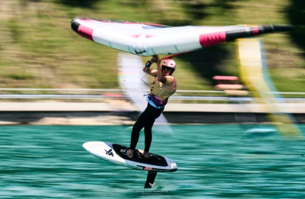 Victor Glatre, champion du monde des moins de 17 ans, navigue sur une planche à hydrofoil tout en tenant une aile gonflable à la main, glissant à toute vitesse sur les eaux bleu-vert de Silvaplana, avec un arrière-plan flou qui suggère un mouvement rapide.